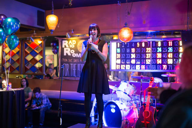 Woman in a black dress speaking into a microphone at a lively event with balloons and a drum set, highlighting community engagement for My Voice Music during Love Bomb 2017.
